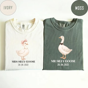 Peut inclure: Deux t-shirts, un ivoire et un vert mousse, chacun avec une illustration d'oie. Le t-shirt ivoire pr&eacute;sente une oie avec un n&oelig;ud rose et le texte "MRS SILLY GOOSE 20.08.2022". Le t-shirt vert affiche une oie avec un n&oelig;ud noir et le texte "MR SILLY GOOSE 20.08.2022".