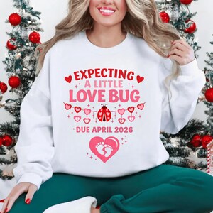 Op de afbeelding: Witte sweatshirt met de tekst "EXPECTING A LITTLE LOVE BUG" in roze en rood. Het ontwerp bevat harten, een lieveheersbeestje en de tekst "DUE APRIL 2026". De sweatshirt is een zwangerschapsaankondiging.