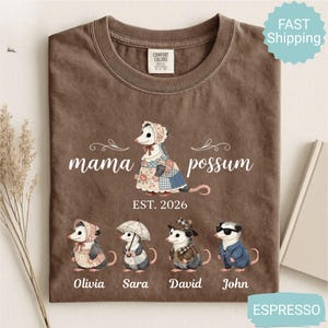 May include: Espresso-colored t-shirt featuring a "mama possum" design with a possum illustration and the text "EST. 2026". Below are illustrations of possums named Olivia, Sara, David, and John. The shirt has a crew neck and short sleeves.