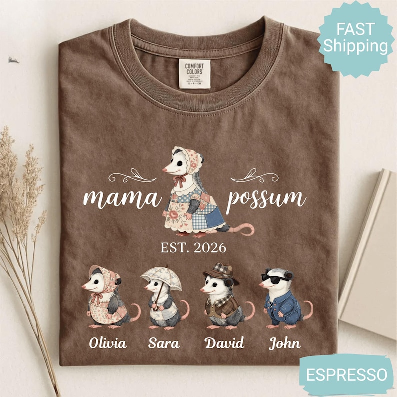 May include: Espresso-colored t-shirt featuring a "mama possum" design with a possum illustration and the text "EST. 2026". Below are illustrations of possums named Olivia, Sara, David, and John. The shirt has a crew neck and short sleeves.