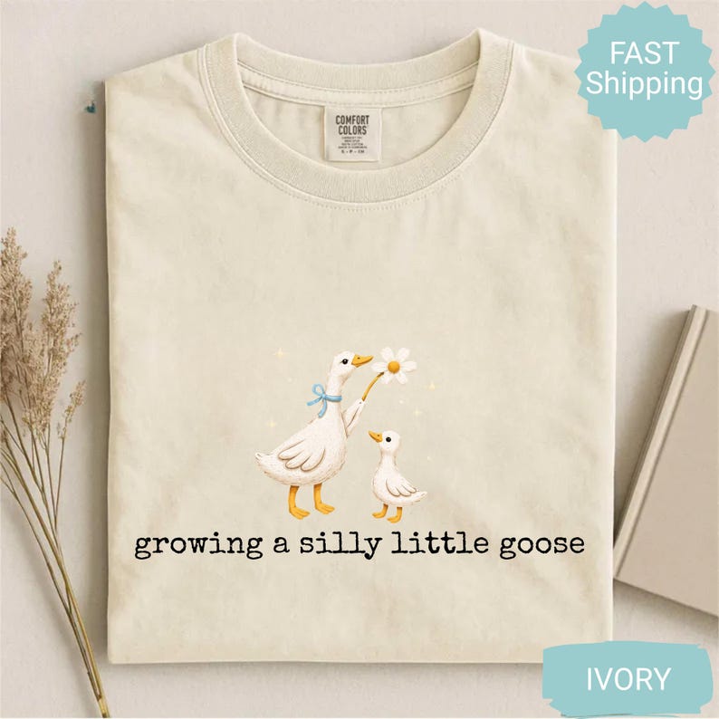 Może przedstawiać: Kremowa koszulka z zabawną ilustracją dw&oacute;ch gęsi. Jedna gęś trzyma stokrotkę, a pod spodem wydrukowano napis "growing a silly little goose". Koszulka ma okrągły dekolt i metkę "Comfort Colors".