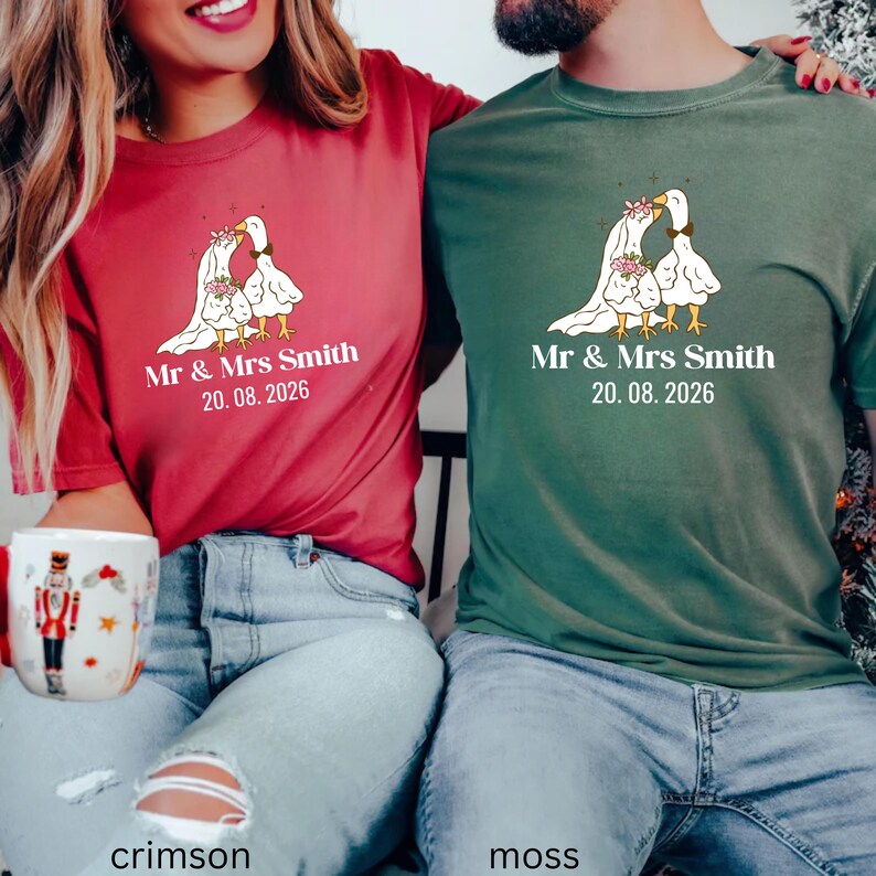 May include: Crimson and moss green t-shirts featuring a wedding-themed design. The graphic shows two geese with floral accents, a veil, and the text "Mr & Mrs Smith 20.08.2026". The shirts are casual and suitable for couples.
