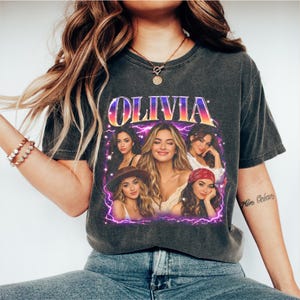May include: Dark grey t-shirt with the name "OLIVIA" in a retro font and a portrait of several women. The graphic is framed by a purple lightning bolt design. The shirt has a vintage style.
