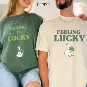 May include: Two people wearing matching t-shirts. One is green and says "Feeling extra Lucky" with a goose holding a shamrock. The other is cream-coloured and says "Feeling Lucky" with a goose holding a shamrock.