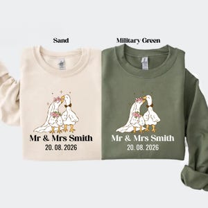 May include: Two sweatshirts, one sand-colored and one military green, each featuring a wedding-themed design. The design includes two geese in wedding attire, the text "Mr & Mrs Smith" and the date "20.08.2026".