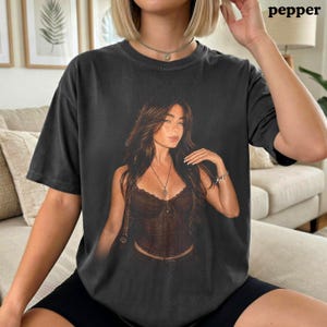 Puede incluir: Una camiseta gris carb&oacute;n de manga corta con un gran estampado gr&aacute;fico centrado de una persona con cabello largo y casta&ntilde;o que lleva un top tipo cors&eacute; marr&oacute;n oscuro y un collar. La camiseta tiene cuello redondo y un ajuste relajado.