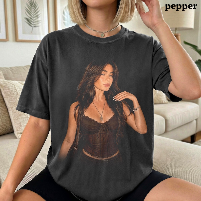 Puede incluir: Una camiseta gris carb&oacute;n de manga corta con un gran estampado gr&aacute;fico centrado de una persona con cabello largo y casta&ntilde;o que lleva un top tipo cors&eacute; marr&oacute;n oscuro y un collar. La camiseta tiene cuello redondo y un ajuste relajado.