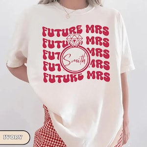 K&ouml;nnte beinhalten: Elfenbeinfarbenes T-Shirt mit rotem Design. Das Design zeigt die Worte "FUTURE MRS" wiederholt, umgeben von einer Diamantring-Grafik mit dem Namen "Smith" darin. Ein l&auml;ssiges, bequemes T-Shirt.