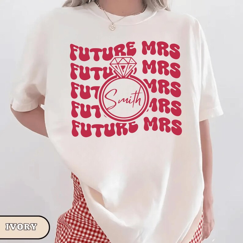 K&ouml;nnte beinhalten: Elfenbeinfarbenes T-Shirt mit rotem Design. Das Design zeigt die Worte "FUTURE MRS" wiederholt, umgeben von einer Diamantring-Grafik mit dem Namen "Smith" darin. Ein l&auml;ssiges, bequemes T-Shirt.