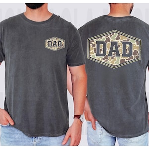 May include: Charcoal gray t-shirt with the word "DAD" in a camouflage design on the front and back. The shirt has a crew neck and short sleeves. The "DAD" design is inside a hexagon shape.