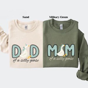 May include: Two sweatshirts, one sand-colored and one military green, each featuring a whimsical design. The sand-colored sweatshirt says "DAD of a silly goose" and the military green sweatshirt says "MOM of a silly goose". Both designs include a goose.