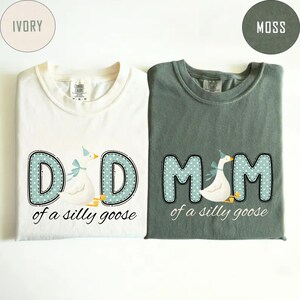 May include: Two t-shirts, one ivory and one moss green, with matching designs. Each shirt features the words "DAD" and "MOM" in a blue polka-dot font, with a cartoon goose wearing a party hat. The text "of a silly goose" is below.