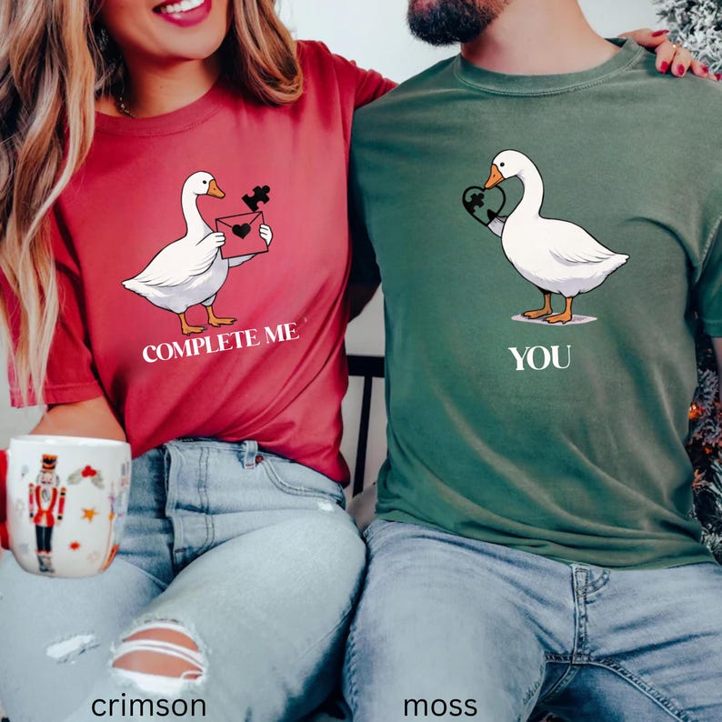 May include: Crimson and moss green t-shirts with goose graphics. The crimson shirt says "Complete Me" and features a goose holding a puzzle piece and an envelope. The moss green shirt says "You" and has a goose holding a heart-shaped puzzle piece.