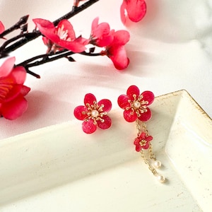 以下が含まれることがあります： 金色のアクセントとイミテーションパールが付いた赤い花のイヤリングのペア。片方のイヤリングはスタッドで、もう片方は追加の花とパールが付いたぶら下がりのチェーンが付いています。イヤリングは、赤い花の枝のある白い背景に設定されています。