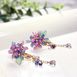 May include: Two floral hair clips with purple and blue petals and pearl accents. The gold-coloured clips have a light green leaf and a chain with beads and a teardrop charm. The clips are suitable for various hairstyles.