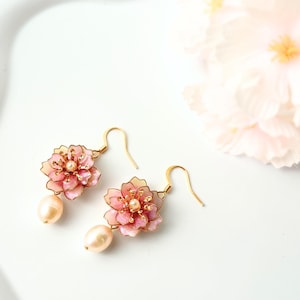 以下が含まれることがあります： ピンクの花の形をしたペンダントとパールのアクセントが付いた、金色のドロップイヤリングのペア。各イヤリングには、洋梨型の薄いピーチ色のパールが付いています。イヤリングは白い背景に配置されています。