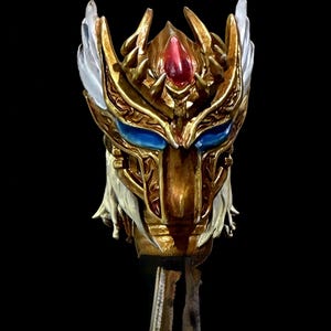 May include: A detailed, golden mask with blue eyes and a red gem at the forehead. The mask features intricate designs and white feathered accents. The mask is mounted on a stand against a black background.