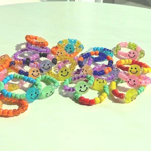 Pode incluir: Uma coleção de anéis coloridos de contas com desenhos de rostos sorridentes. Os anéis são de várias cores, incluindo laranja, rosa, azul, verde e amarelo. Os anéis são feitos de pequenas contas redondas e estão dispostos sobre uma superfície branca.