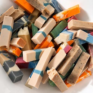 May include: A collection of rectangular soap bars in various colours and textures, including solid beige, bright orange, dark grey, and blue-swirled patterns, piled together in a white basin with a visible metal drain at the top right corner.
