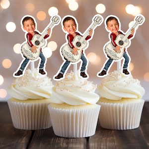 May include: Three cupcakes with white frosting and pearl sprinkles, each topped with a guitar-playing boy figurine. The boy wears a red jacket, blue jeans, and brown shoes. The guitar is white with decorative detailing.