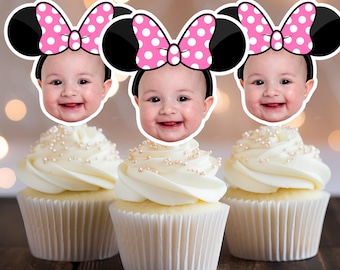 Op de afbeelding: Drie cupcakes met witte glazuur en parelkleurige sprinkles. Elke cupcake is bekroond met een babygezicht met een Minnie Mouse oren hoofdband met een roze strik en witte stippen. De achtergrond is vervaagd met bokeh-lichten.