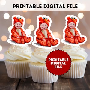 May include: Three cupcakes topped with baby-themed toppers. Each topper features a baby in a red costume. The cupcakes have white frosting and are decorated with small, silver sprinkles. The text "PRINTABLE DIGITAL FILE" is displayed.