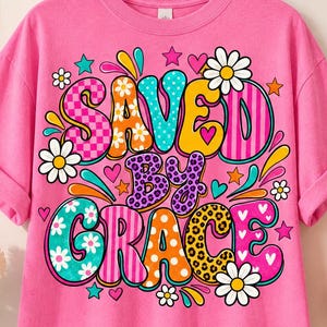 May include: A bright pink short-sleeve t-shirt featuring a colourful graphic print with the text "SAVED BY GRACE" in large, multi-patterned bubble letters. The design includes various floral motifs, stars, hearts, and abstract shapes in vibrant colours.