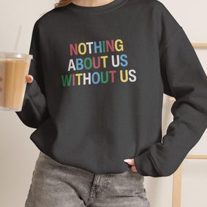 Könnte beinhalten: Ein schwarzes Sweatshirt mit Rundhalsausschnitt und dem Text "NOTHING ABOUT US WITHOUT US" in einer mehrfarbigen Schriftart. Die Buchstaben sind in Rot, Gelb, Blau und Grün gehalten und heben sich deutlich vom dunklen Stoff des langärmeligen Oberteils ab.