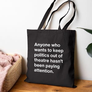 May include: A black tote bag with two shoulder straps featuring white text that reads: Anyone who wants to keep politics out of theatre hasn't been paying attention. The bag is constructed from a solid black fabric with bold, sans-serif lettering.
