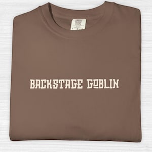 Puede incluir: Una camiseta marrón de cuello redondo con el texto BACKSTAGE GOBLIN impreso en una fuente de estilo bloque de color crema en el pecho. La prenda incluye una etiqueta de la marca Comfort Colors en el cuello interior, indicando la talla grande.