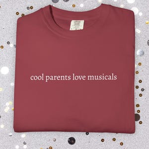 Puede incluir: Una camiseta de cuello redondo de color rojo oscuro con el texto blanco "cool parents love musicals" impreso en el centro del pecho. La camiseta tiene un cuello estándar y una etiqueta de marca visible en el interior del escote.