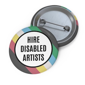 May include: A round pin-back button featuring a white center circle with the black text HIRE DISABLED ARTISTS. The border of the button displays a diagonal striped pattern in colors including yellow, red, black, green, and light blue. The back is metal.