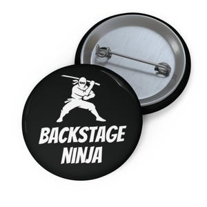 May include: A round pin-back button featuring a black background with a white graphic of a ninja holding a sword above their head. Below the ninja, the text BACKSTAGE NINJA is printed in bold white capital letters. The back shows a metal pin mechanism.