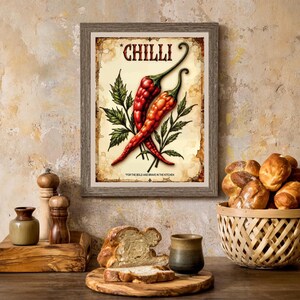 K&ouml;nnte beinhalten: Ein gerahmter Druck mit zwei roten Chilischoten und gr&uuml;nen Bl&auml;ttern, dem Wort "CHILLI" oben und Text unten. Der Druck befindet sich an einer Wand, mit Brot und einem Korb auf einem Holztisch im Vordergrund.