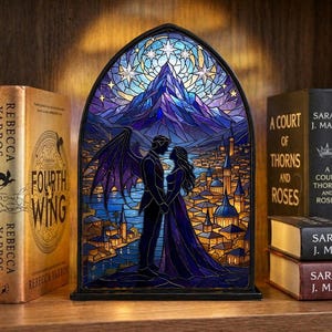 Peut inclure: Une veilleuse de style vitrail représentant un couple devant une scène de montagne et de ville. La lumière a un cadre noir et est entourée de livres sur une étagère en bois. La palette de couleurs comprend des bleus, des violets et de l'or.
