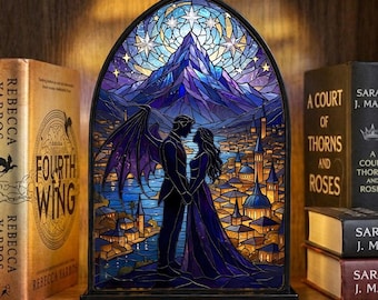May include: A stained glass-style night light featuring a couple holding hands in front of a mountain and city scene. The light has a black frame and is surrounded by books on a wooden shelf. The color palette includes blues, purples, and gold.