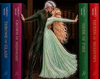 May include: A clear acrylic standee featuring a male figure with silver hair in a dark green coat and a female figure with long blonde hair in a light green gown, posed in a dance. The standee is positioned between two rows of hardcover books.