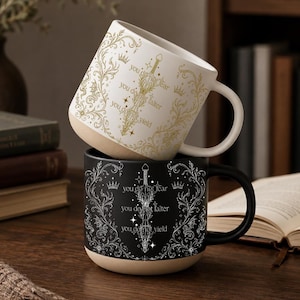 May include: Two ceramic mugs, one white and one black, stacked on a wooden surface. The white mug has gold floral and crown designs with the text "you do not fear, you do not falter, you do not yield." The black mug has similar silver designs and text.