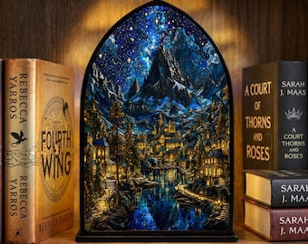 May include: A stained glass window depicting a nighttime mountain scene with a starry sky, a river, and a town with lit windows. The window is arched and sits on a shelf with books.