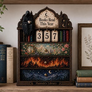 Puede incluir: Un organizador de estantería de madera marrón oscuro con un letrero que dice Books Read This Year y tres tarjetas numéricas colgantes que muestran 357. Una figura negra con capucha se sienta encima. Tres cajones muestran escenas florales, de fuego y de cielo nocturno. Hannah's Library está impreso en la base.