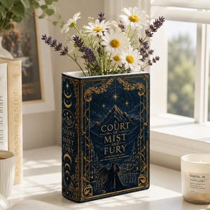 May include: A rectangular ceramic vase shaped like a book with a dark blue cover featuring gold celestial patterns, mountain illustrations, and the text A Court of Mist and Fury. The vase holds a bouquet of white daisies and purple lavender stems.