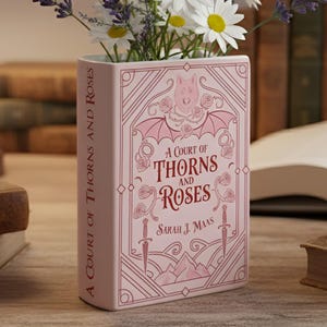 Puede incluir: Un jarrón de cerámica rosa con forma de libro, con el título "A Court of Thorns and Roses" e ilustraciones florales. El jarrón contiene margaritas blancas y lavanda morada. Está sobre una superficie de madera, con libros y una lámpara.