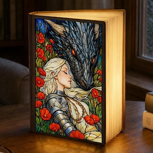 Puede incluir: Lámpara decorativa con forma de libro con una ilustración colorida de una mujer y un dragón. La ilustración presenta a una mujer con pelo largo rubio, amapolas rojas y un dragón con ojos rojos. La lámpara está abierta y las páginas están iluminadas.
