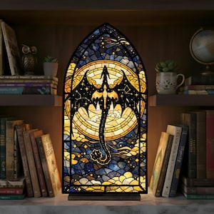 May include: A stained glass lamp featuring a dragon design. The lamp has a black frame and a colorful mosaic of blues, yellows, and oranges. The dragon is black with outstretched wings. The lamp is on a wooden base and is illuminated from within.