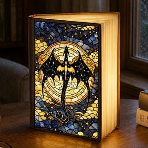 Puede incluir: Lámpara decorativa con forma de libro con un diseño de vidriera que presenta una silueta de dragón negro. La lámpara emite un brillo cálido, iluminando las páginas y la ilustración del dragón. El fondo es una mezcla de azul, amarillo y dorado.