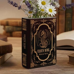 May include: A decorative vase shaped like a book, featuring the title "Fourth Wing" and a dragon illustration. The vase is filled with white daisies and purple lavender. The vase is dark brown with gold accents, and is sitting on a wooden surface.