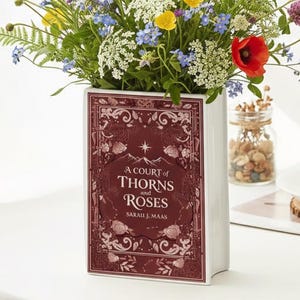 May include: A book-shaped white ceramic vase displaying the title "A Court of Thorns and Roses". The vase is filled with a colourful arrangement of wildflowers, including red poppies, yellow buttercups, and blue forget-me-nots.