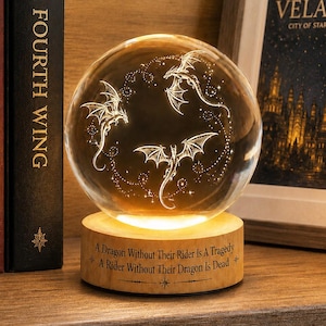 May include: A clear glass sphere lamp featuring three etched flying dragons, illuminated from a light-coloured wooden base. The base is engraved with the text: A Dragon Without Their Rider Is A Tragedy, A Rider Without Their Dragon Is Dead.