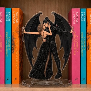May include: A clear acrylic standee featuring a couple with large black wings, the person on the left wears a long black glittery dress with a high slit, and the person on the right wears black armour, positioned between two rows of book spines.