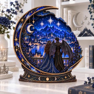 May include: A crescent moon-shaped decorative display featuring a backlit night scene of a fantasy city with mountains and stars. Two figures stand on a balcony, one with long hair in a blue gown and one with dark wings in a black coat.